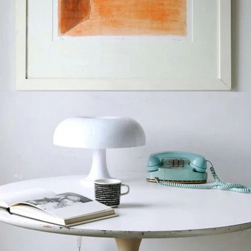 Weiße Tischlampe, Retro-Telefon, Kaffeetasse auf rundem Tisch vor Kunstwerk an weißer Wand.