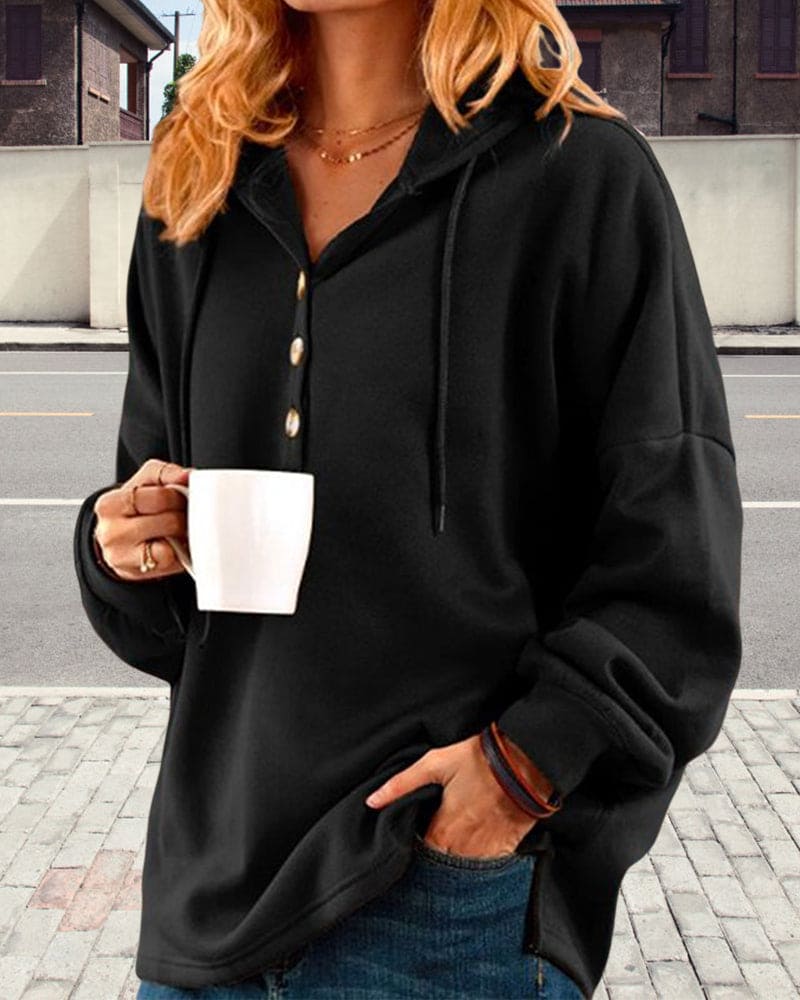 Frau in schwarzem Kapuzenpullover mit Knöpfen hält weiße Tasse, lässige Mode, Straße im Hintergrund.
