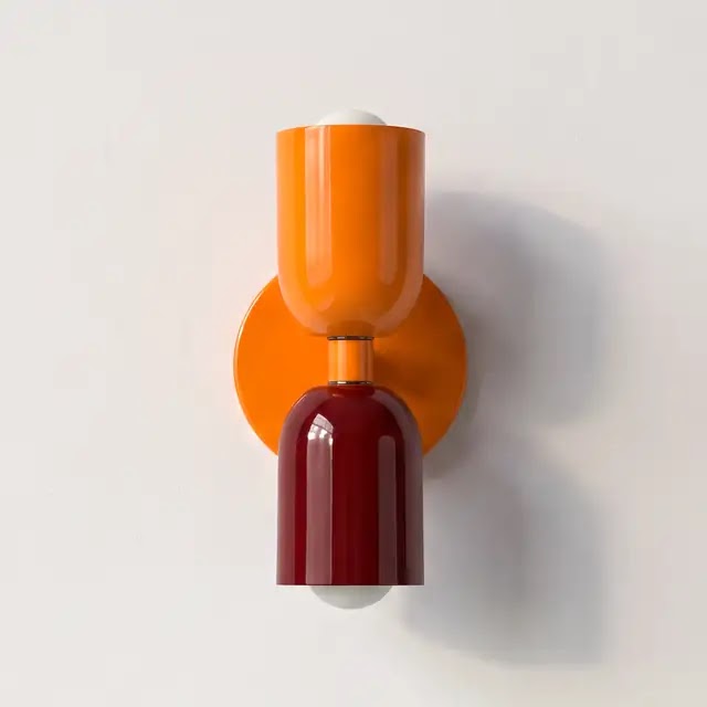 Vägglampa med orange övre skärm och orange väggplatta samt blank vinröd nedre skärm, uppsatt på en ljus vägg. 25