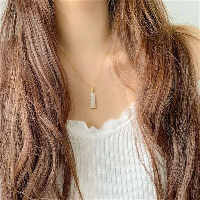 Frau mit langem Haar trägt goldene Halskette mit rechteckigem Anhänger, weißes Oberteil.
