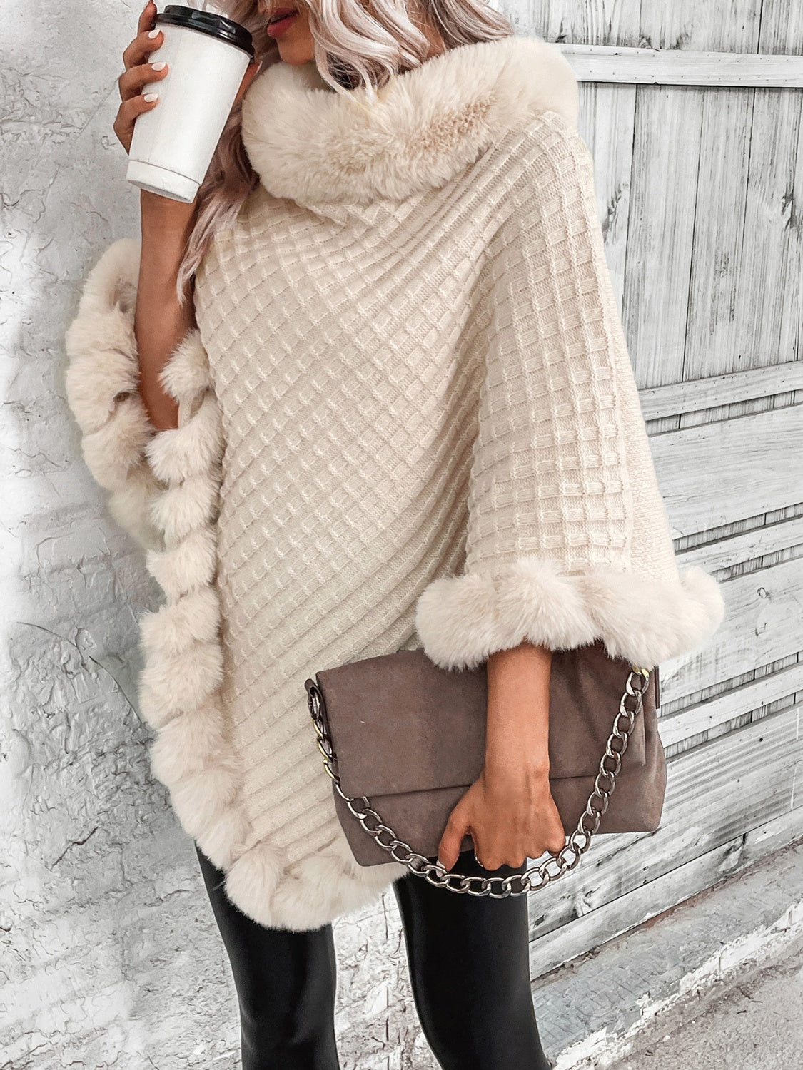 Frau in cremefarbenem Strickponcho mit Kunstpelz, hält braune Tasche und Kaffeebecher.