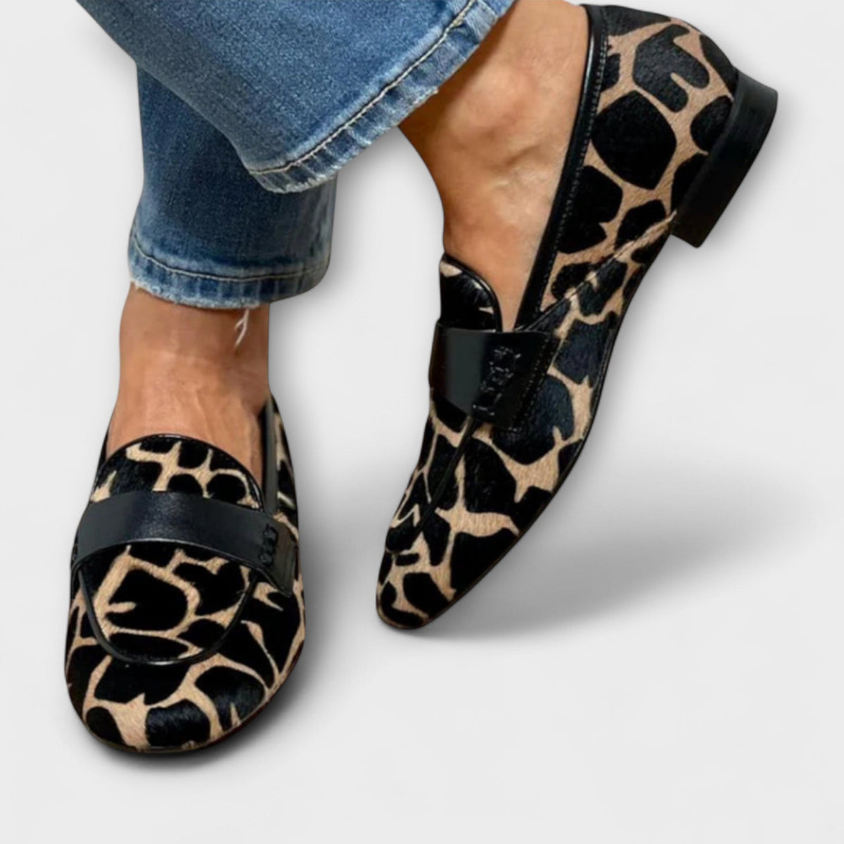 Ett par loafers med svart läderrem och kantlist, sedda uppifrån och från sidan med platt svart klack, burna med jeans. 3
