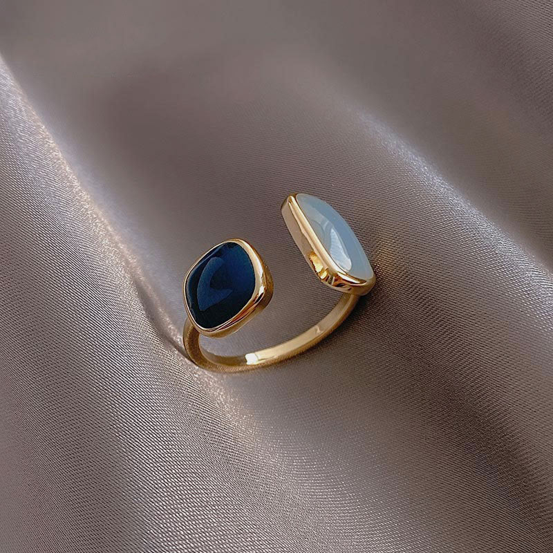 Goldener offener Ring mit blauem und weißem Stein, elegantes Schmuckdesign auf Seidenstoff.