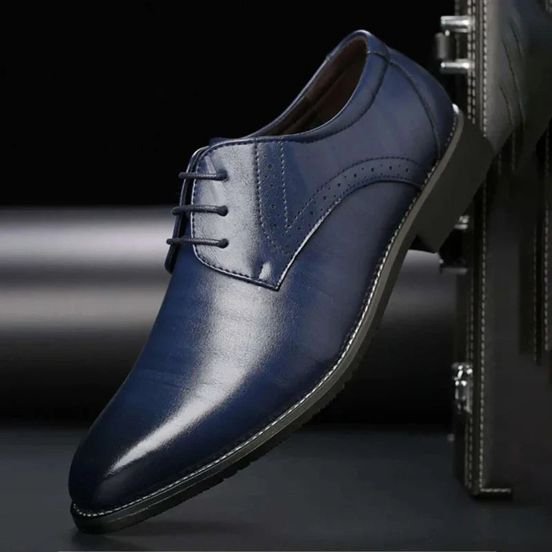 Blå blank läderskor med snörning och brogue-perforering, fotograferad snett framifrån mot en mörk bakgrund och portfölj. 5