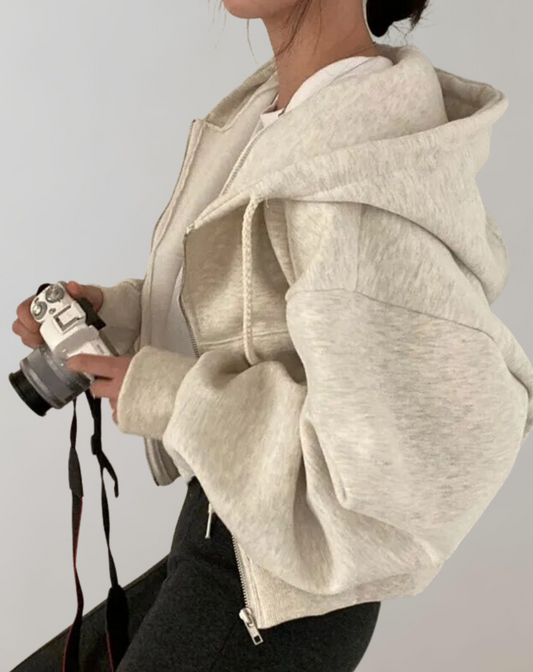 Modell i ljus gråbeige hoodie sedd från sidan, med stor huva och vida ärmar, håller en kamera i händerna. 2