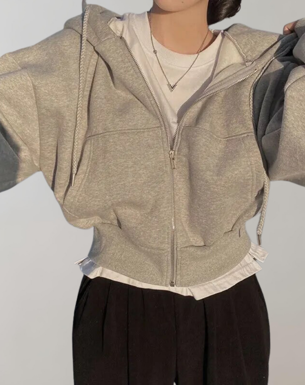 Modell i ljusgrå hoodie med dragkedja och ribbad nederkant, över vit tröja som sticker fram, med svarta byxor och armarna upplyfta. 5
