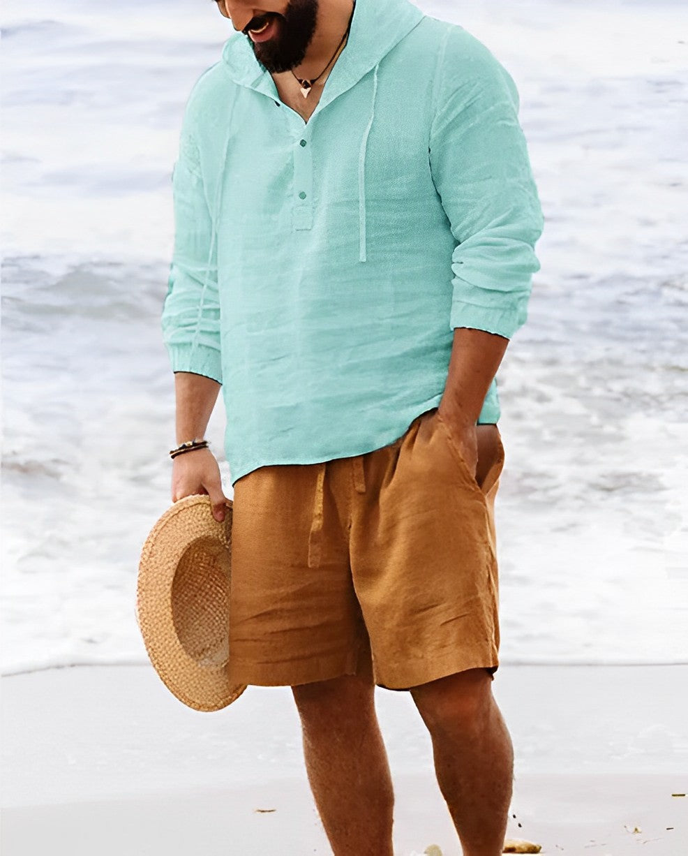 Mann am Strand in türkisfarbenem Leinenhemd und braunen Shorts, hält Strohhut.