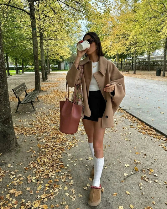 Frau im Herbstoutfit mit beigem Mantel, schwarzem Rock, rosa Tasche, trinkt Kaffee im Park.