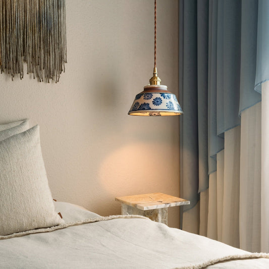 Tänd lampa med blå blommönstrad keramikskärm hänger vid en säng med beige textilier, ett marmorbord och blå gardiner. 2
