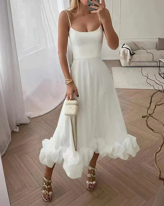 Frau in weißem Sommerkleid mit Rüschen, goldenen Sandalen und weißer Handtasche im Wohnzimmer.