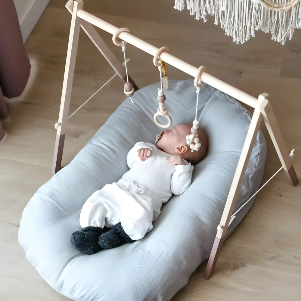 Schlafendes Baby auf grauem Kissen unter Holzspielbogen mit hängendem Spielzeug.