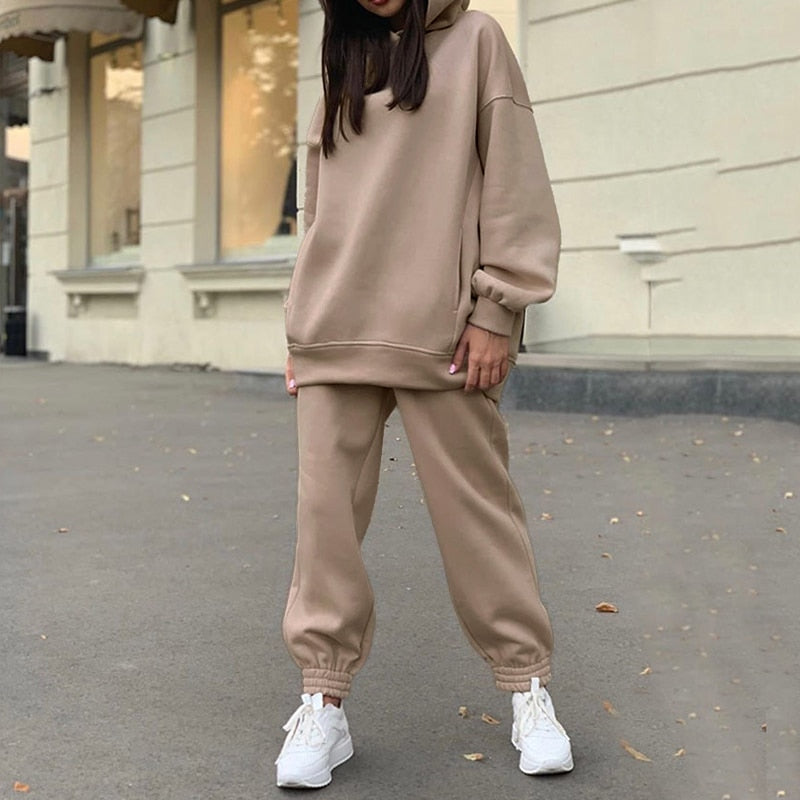 Frau in beigem Jogginganzug, Kapuzenpullover und Jogginghose, weiße Sneaker, urbaner Hintergrund.