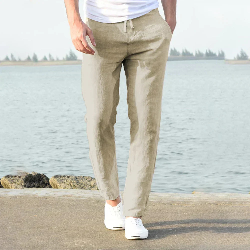Mann in beigen Leinenhose am Wasser, lässiger Sommerstil, weiße Sneaker.