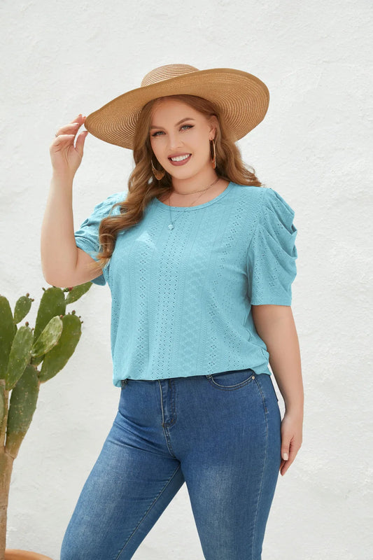 Frau in blauer Spitzenbluse und Jeans, trägt Strohhut, lächelt, vor weißer Wand mit Kaktus.