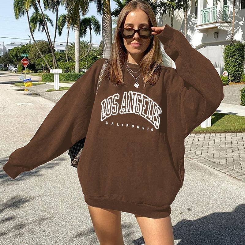 Frau in braunem "Los Angeles" Oversize-Pullover, Sonnenbrille, Straße, Palmen im Hintergrund.