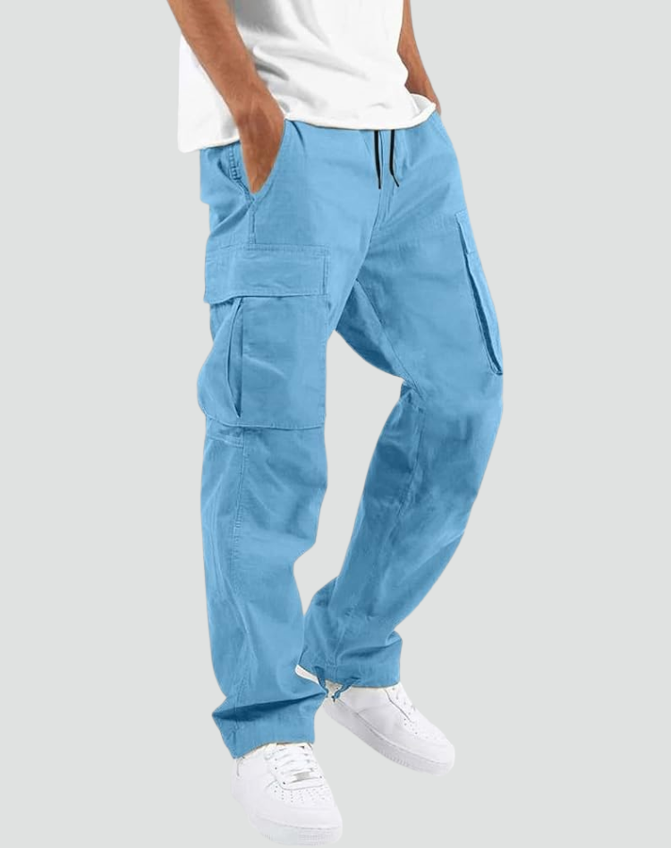 Man bär ljusblå cargobyxor med stora sidfickor och dragsko i midjan, vit t-shirt och vita sneakers, mot grå bakgrund. 14