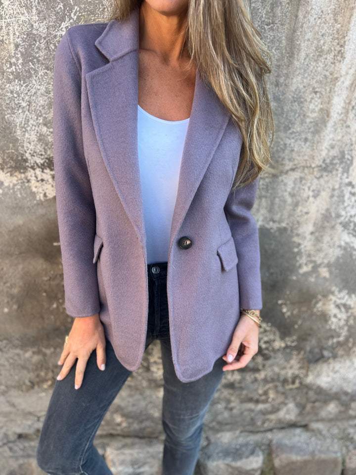 Frau in lila Blazer, weißem Top und Jeans vor Betonwand, modisch, elegant, Herbstmode.