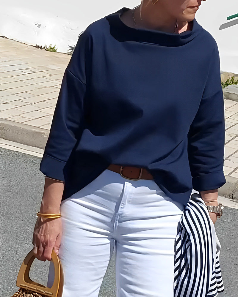 Frau in dunkelblauem Pullover und weißer Hose, hält gestreifte Jacke und Holzhandtasche.