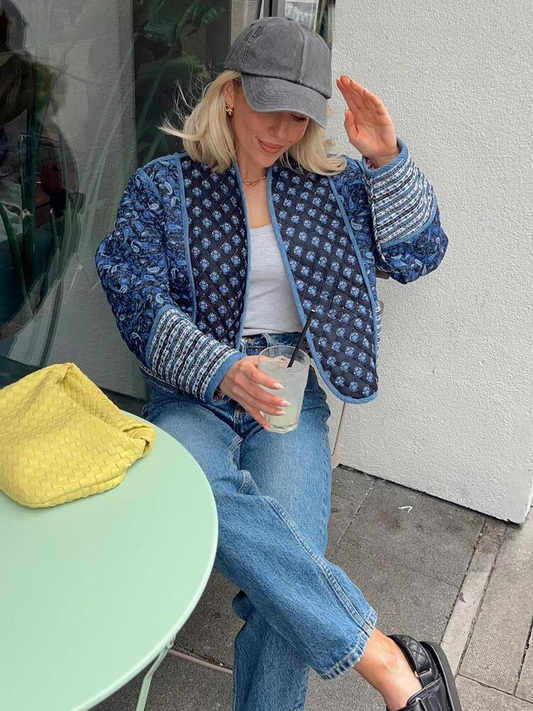 Frau in blauer Steppjacke und Jeans, graue Kappe, gelbe Tasche, sitzt an grünem Tisch draußen.