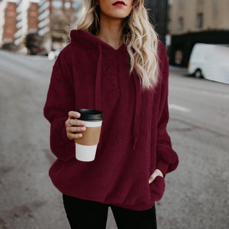 Frau in bordeauxrotem Hoodie hält Kaffeebecher, Stadtstraße im Hintergrund.