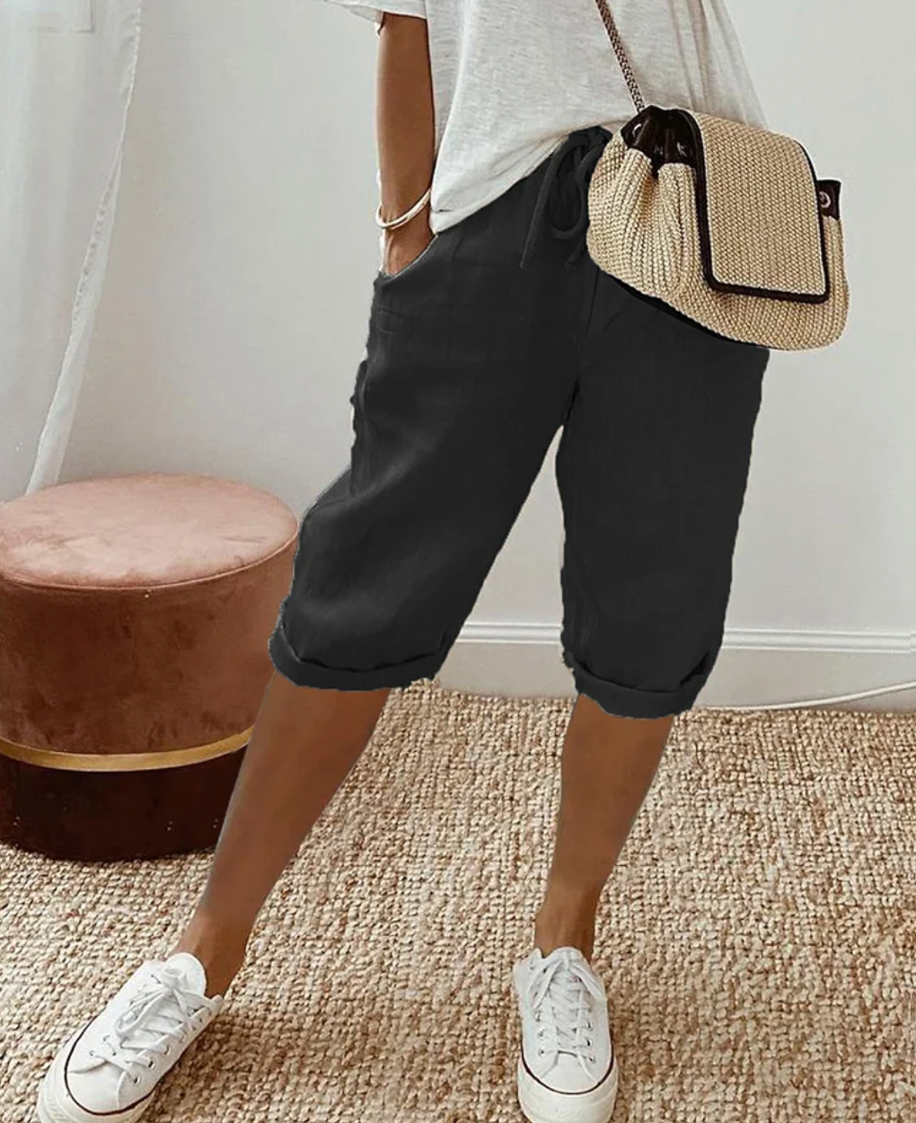 Frau in schwarzer Bermuda-Shorts, weißem T-Shirt, Strohtasche, weiße Sneaker, lässiger Stil.