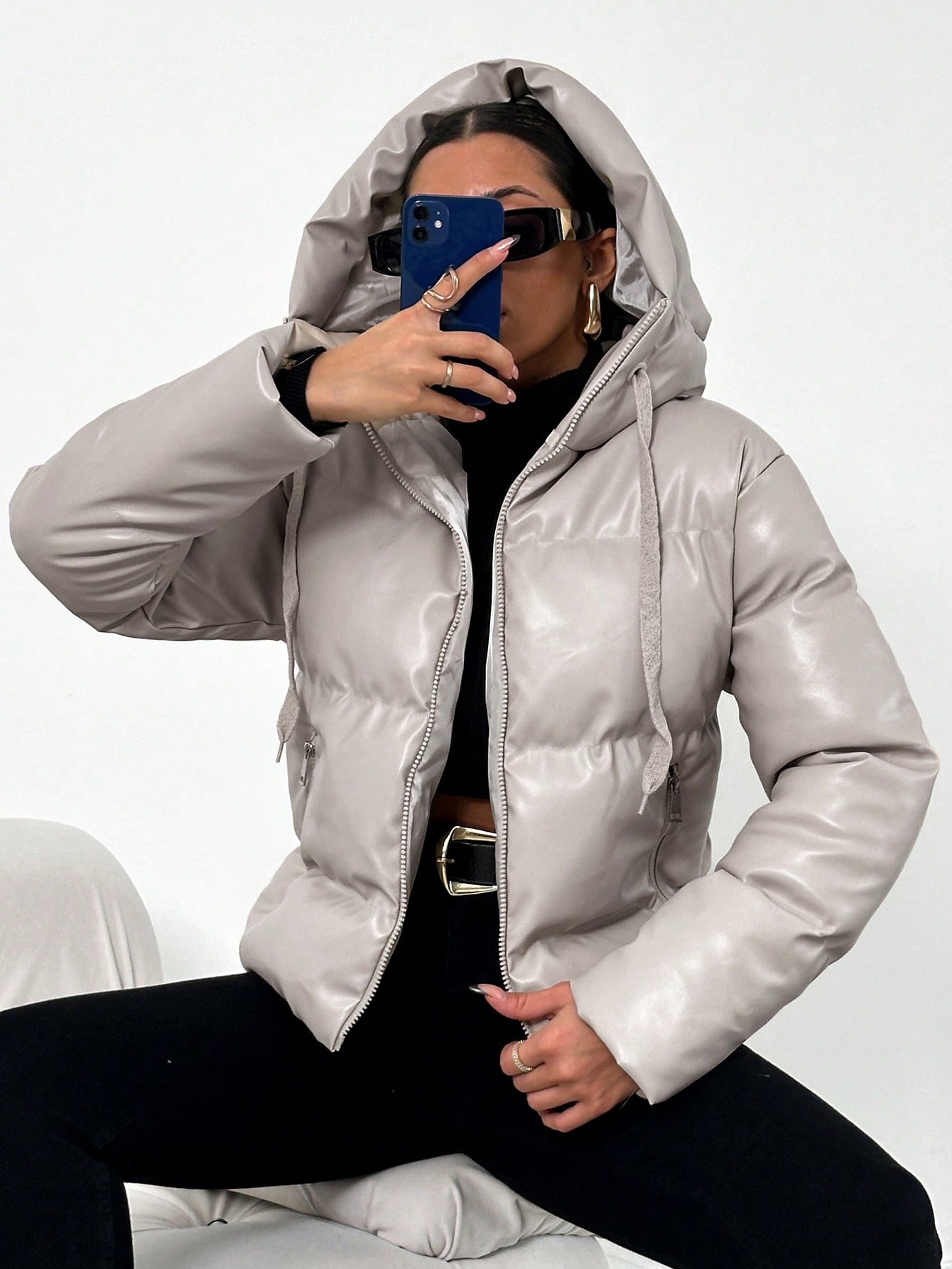 Frau in grauer Daunenjacke mit Kapuze, hält Smartphone, trägt Sonnenbrille, modisch, Winterkleidung.