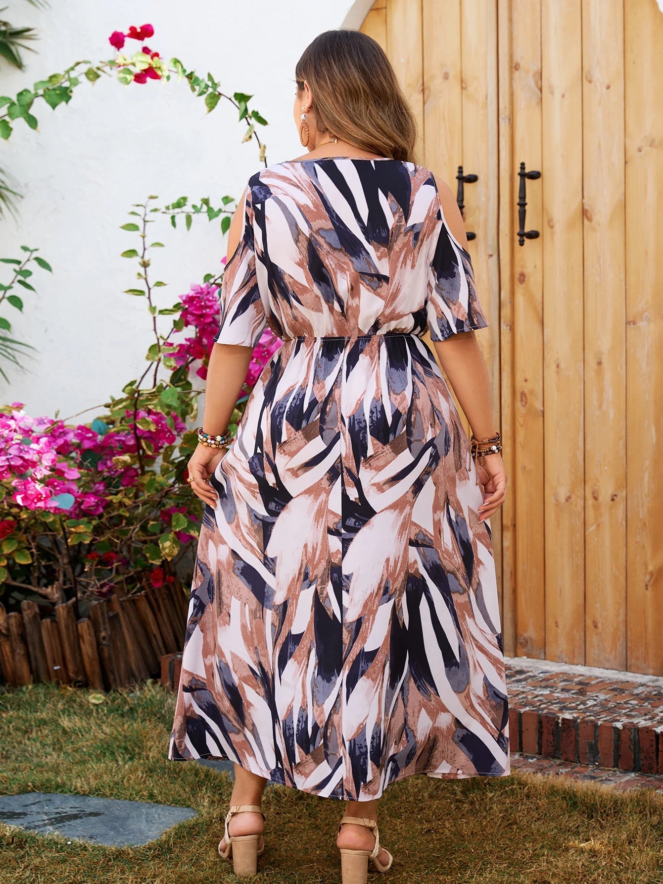 Frau im gemusterten Sommerkleid vor Holztür, Blumen im Hintergrund, modisch, elegant.