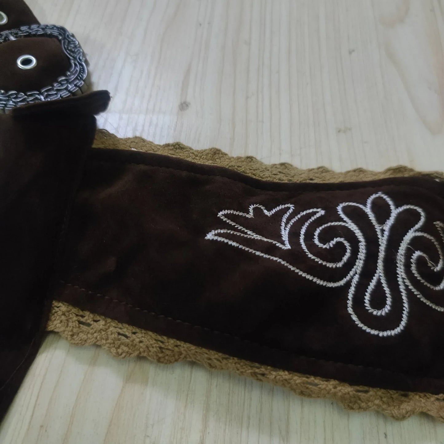 Brauner Ledergürtel mit weißer Stickerei und gehäkeltem Rand auf Holztisch.