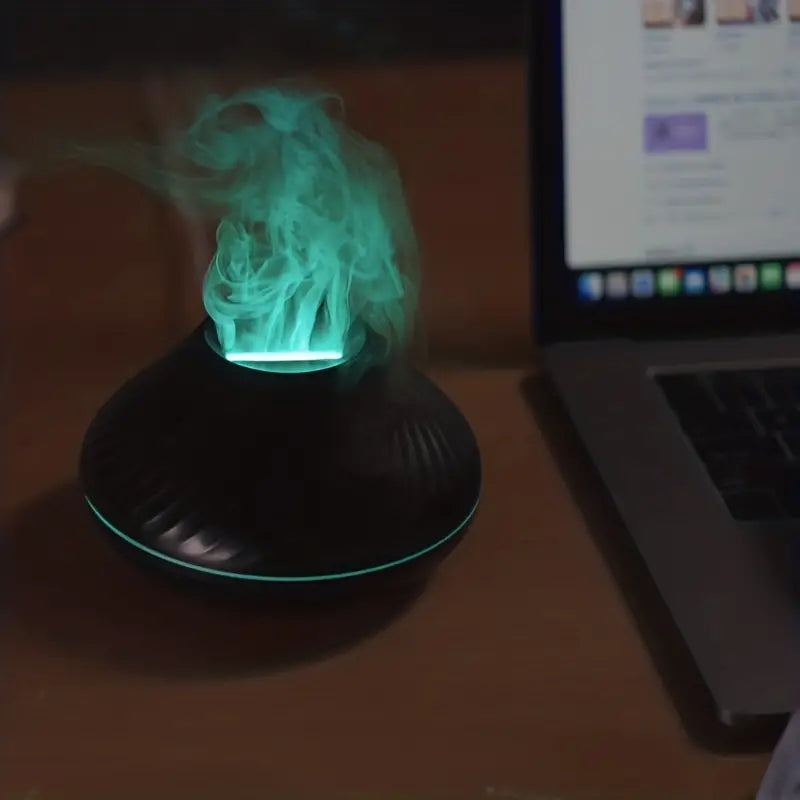 Schwarzer Aromadiffusor mit grünem LED-Licht und Dampf neben Laptop auf Holztisch.