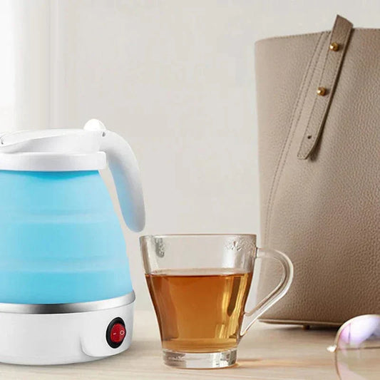 Blauer faltbarer Wasserkocher aus Silikon neben Teetasse und beiger Handtasche auf Holztisch.