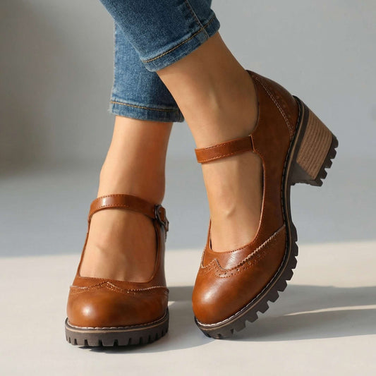 Bruna Mary Jane-skor med brogue-detaljer, justerbar rem och kraftig gummisula, bärs med jeans i studiomiljö. 2