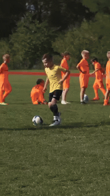 Junge in gelbem Trikot dribbelt Fußball auf grünem Feld, umgeben von Kindern in orangefarbenen Trikots.