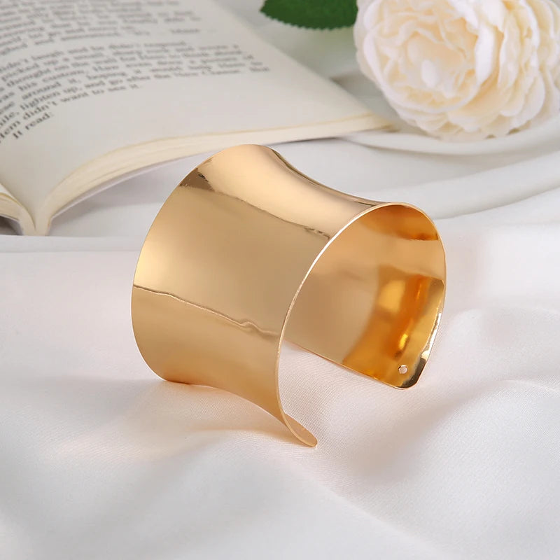 Goldenes Armreif aus Metall auf weißem Stoff, elegantes Schmuckdesign, offenes Buch im Hintergrund.