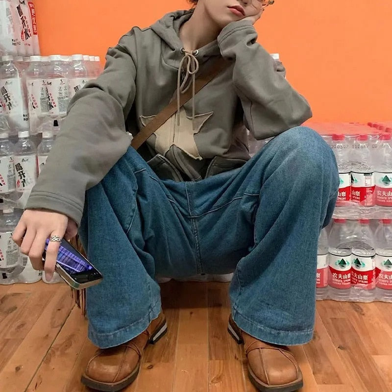 Person in grauem Hoodie und blauer Jeans sitzt vor Wasserflaschen, hält Smartphone, trägt braune Schuhe.