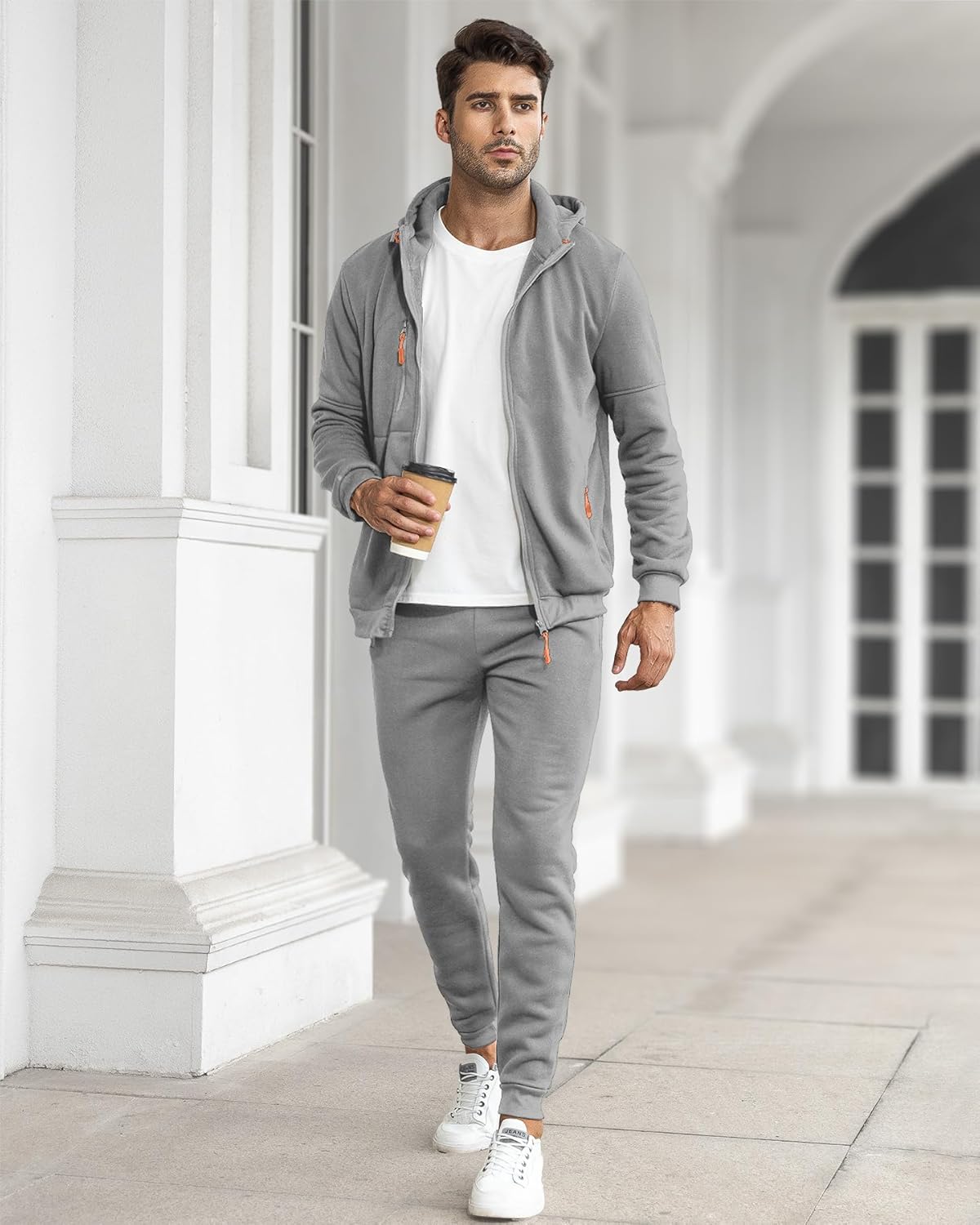Mann im grauen Jogginganzug mit Kapuze, weißem T-Shirt und Sneakers, hält Kaffeebecher.