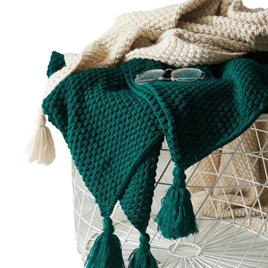 Grüne Strickdecke mit Quasten auf weißem Korb, gemütliche Wohnaccessoires, Brille obenauf.