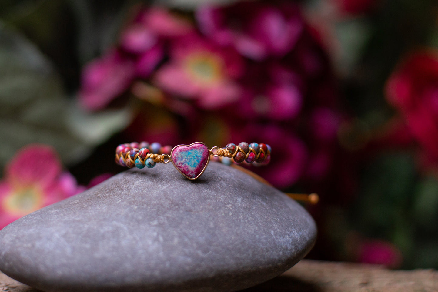 Armband mit Herzanhänger, bunte Perlen, auf Stein, vor rosa Blumen, Schmuck, Geschenkidee.