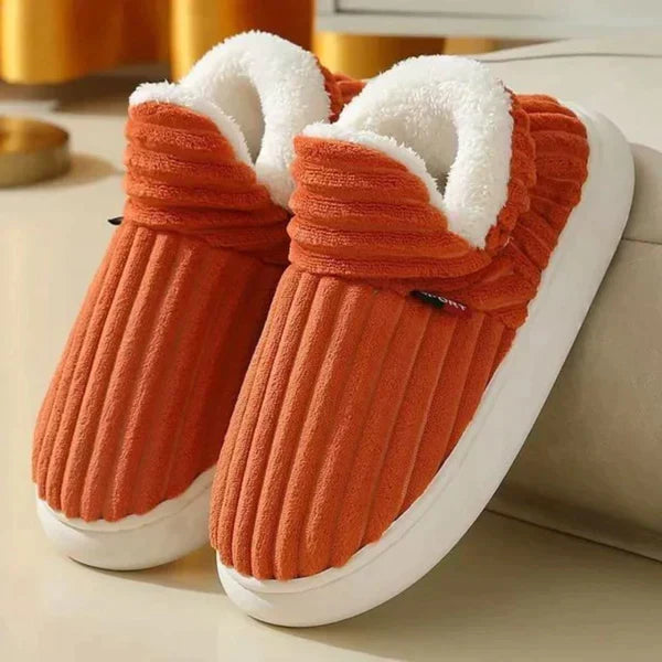 Ett par ribbade plyschtofflor i orangerött med vitt fluffigt foder och tjock vit sula, lutade mot en soffa. 6