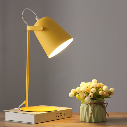Tänd gul bordslampa med konformad skärm på en bok, bredvid en liten grön kruka med vita och gula blommor. 1