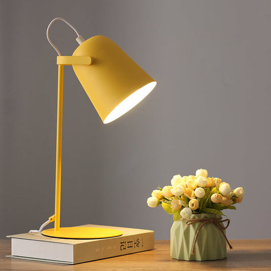 Tänd gul bordslampa med konformad skärm på en bok, bredvid en liten grön kruka med vita och gula blommor. 1