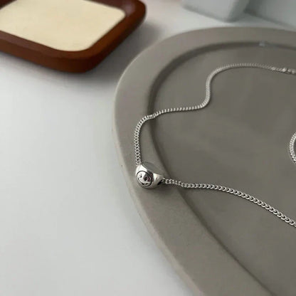 Silberne Halskette mit Smiley-Anhänger auf grauem Hintergrund, minimalistisches Schmuckdesign.