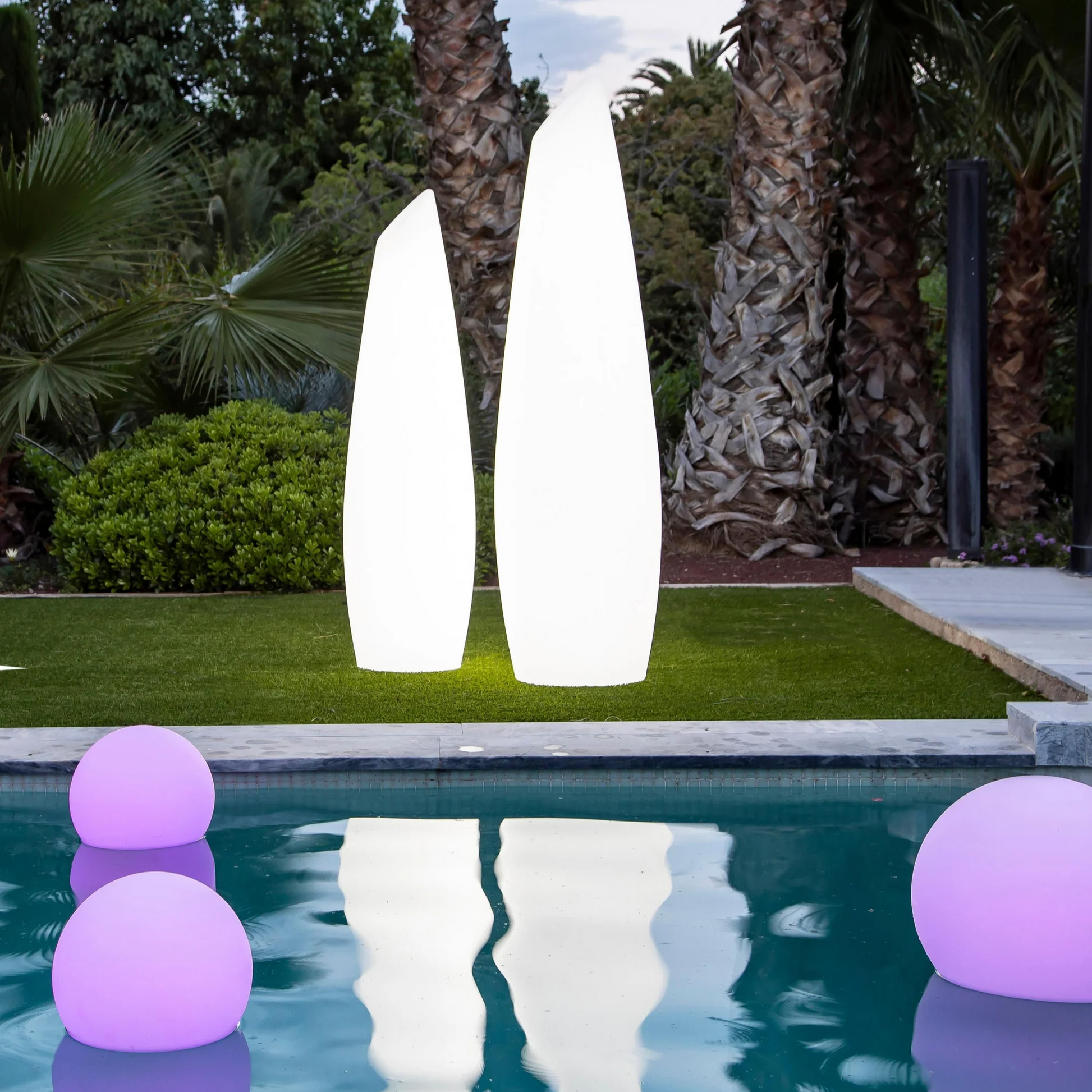Zwei weiße LED-Gartenleuchten neben Pool, umgeben von Palmen und lila Leuchtkugeln.