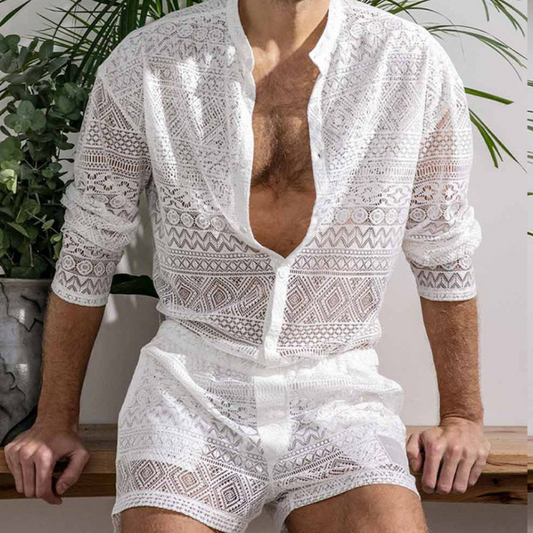 Mann in weißem, transparentem Spitzenhemd und Shorts, sitzt vor Pflanzen.