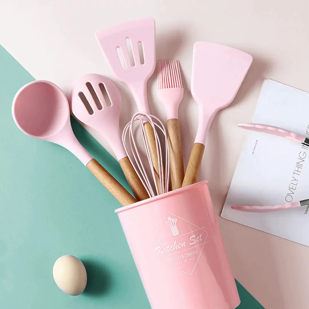 Rosa Küchenutensilien-Set aus Silikon und Holz in einem rosa Behälter auf pastellfarbenem Hintergrund.