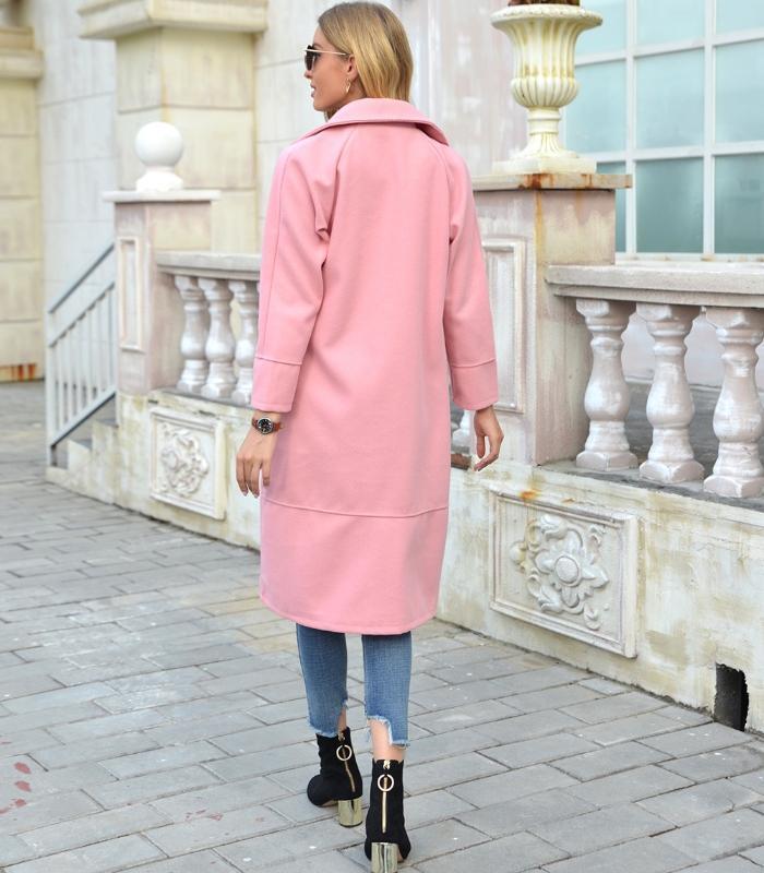 Frau in rosa Mantel, Jeans und Stiefeletten auf Gehweg vor Gebäude.