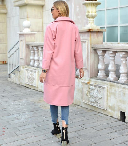 Frau in rosa Mantel, Jeans und Stiefeletten auf Gehweg vor Gebäude.
