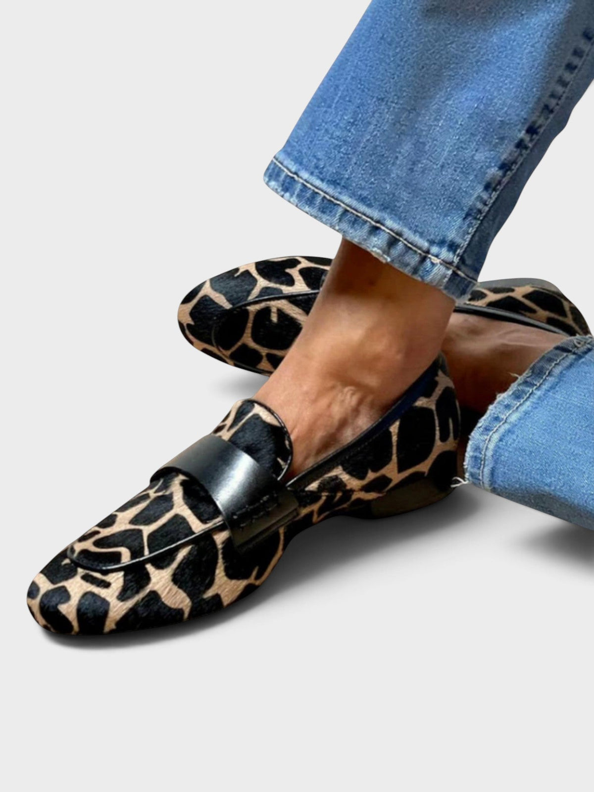 Loafers med svarta och beigea fläckar och bred svart läderrem, burna med jeans, sedda snett framifrån med korsade fötter. 1