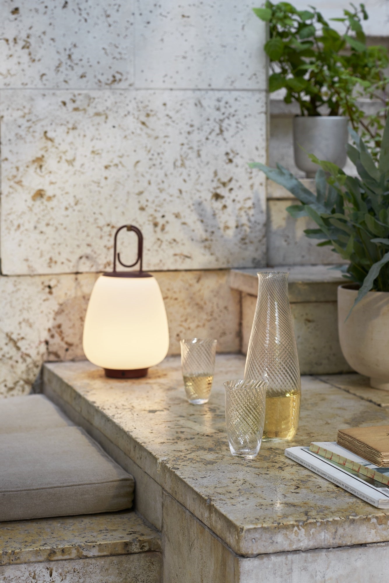 Tragbare LED-Lampe auf Steintreppe, umgeben von Pflanzen und Glaskaraffe, modernes Outdoor-Design.