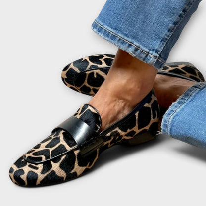 Närbild på de mönstrade loafersen på fötterna, med svart kantlist och läderrem över vristen, mot ljus bakgrund och uppvikta jeans. 2