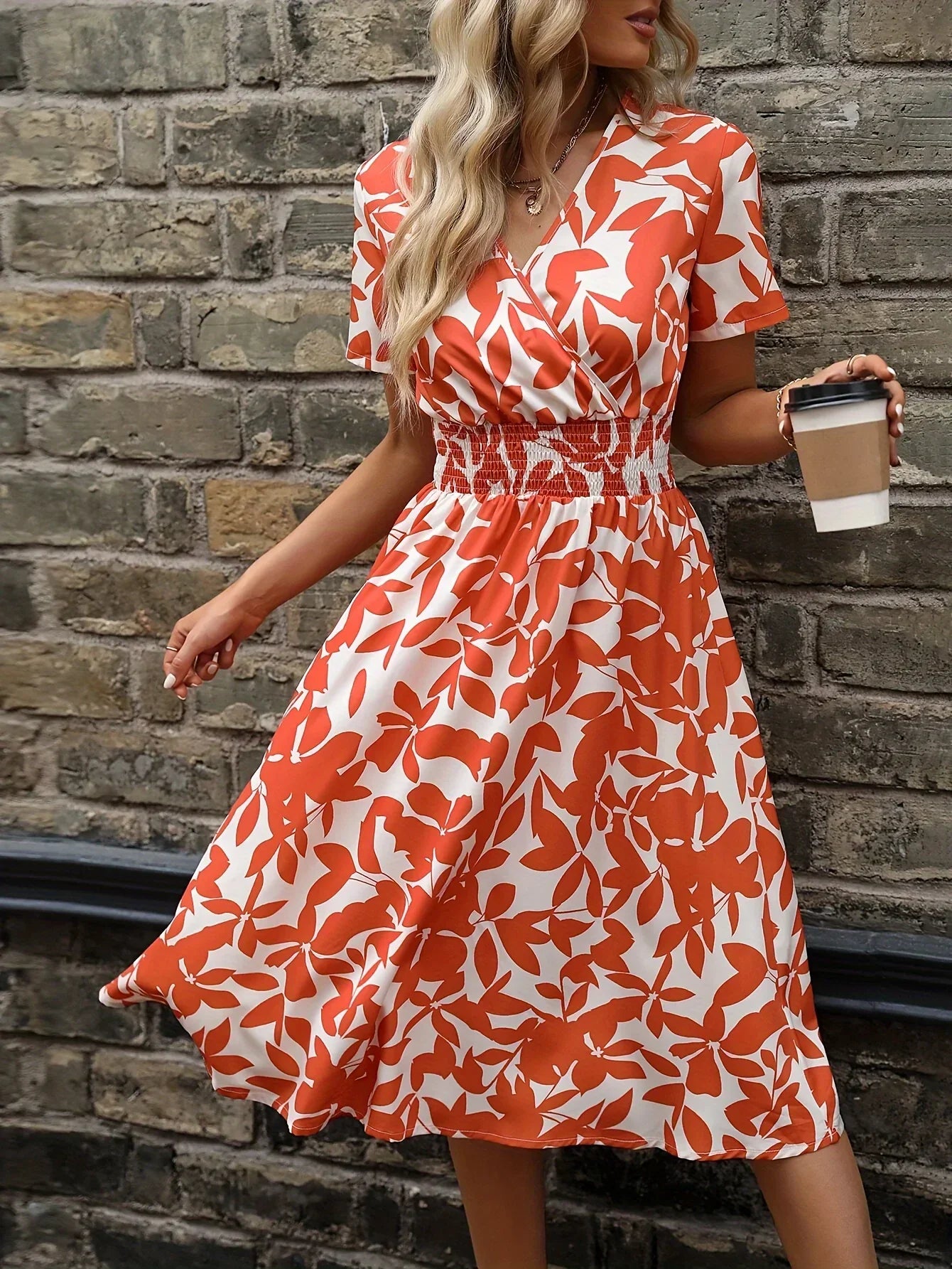 Frau in rotem Sommerkleid mit Blattmuster hält Kaffeebecher vor Backsteinmauer.
